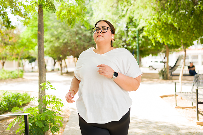 Woman with glasses and smartwatch jogging on a sunny park path surrounded by green trees.