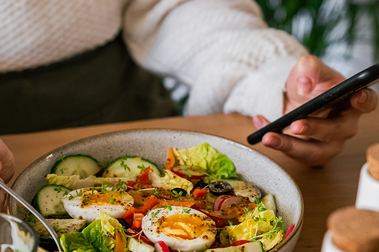 Person taking a photo of a salad bowl with boiled eggs, cucumber slices, lettuce, radishes, and red bell peppers on a wooden table.