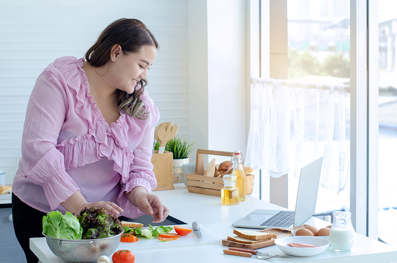 Woman in a pink blouse preparing fresh vegetables on a kitchen counter with bread, eggs, and a laptop nearby.