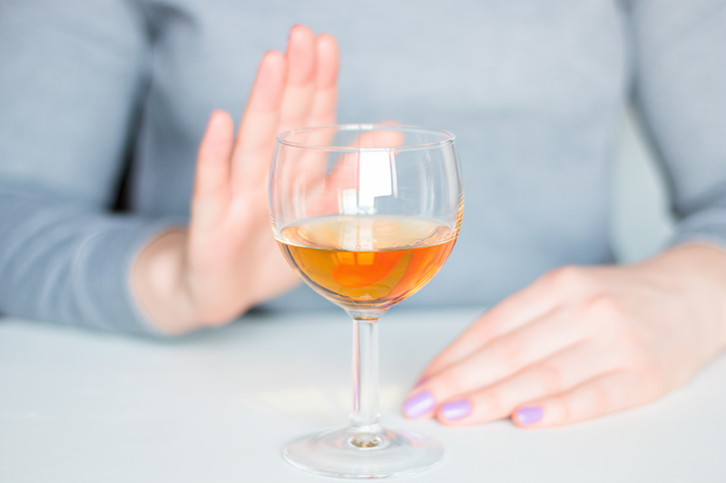 Person with lavender nail polish holding up a hand to refuse a glass of amber-colored alcohol on a white surface.