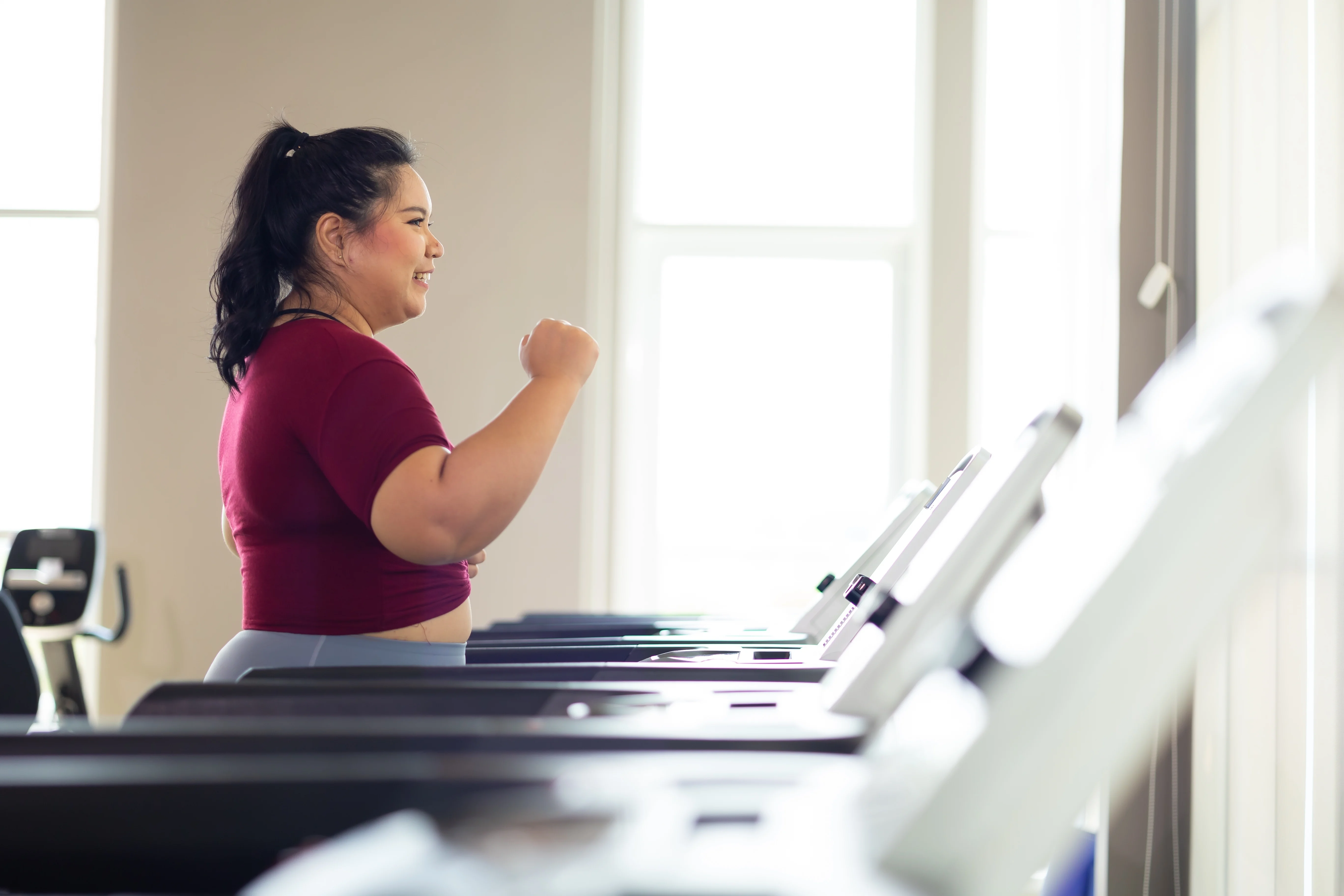 Smiling woman with dark hair tied in a ponytail wearing a red shirt and gray leggings walking on a treadmill in a brightly lit gym.
