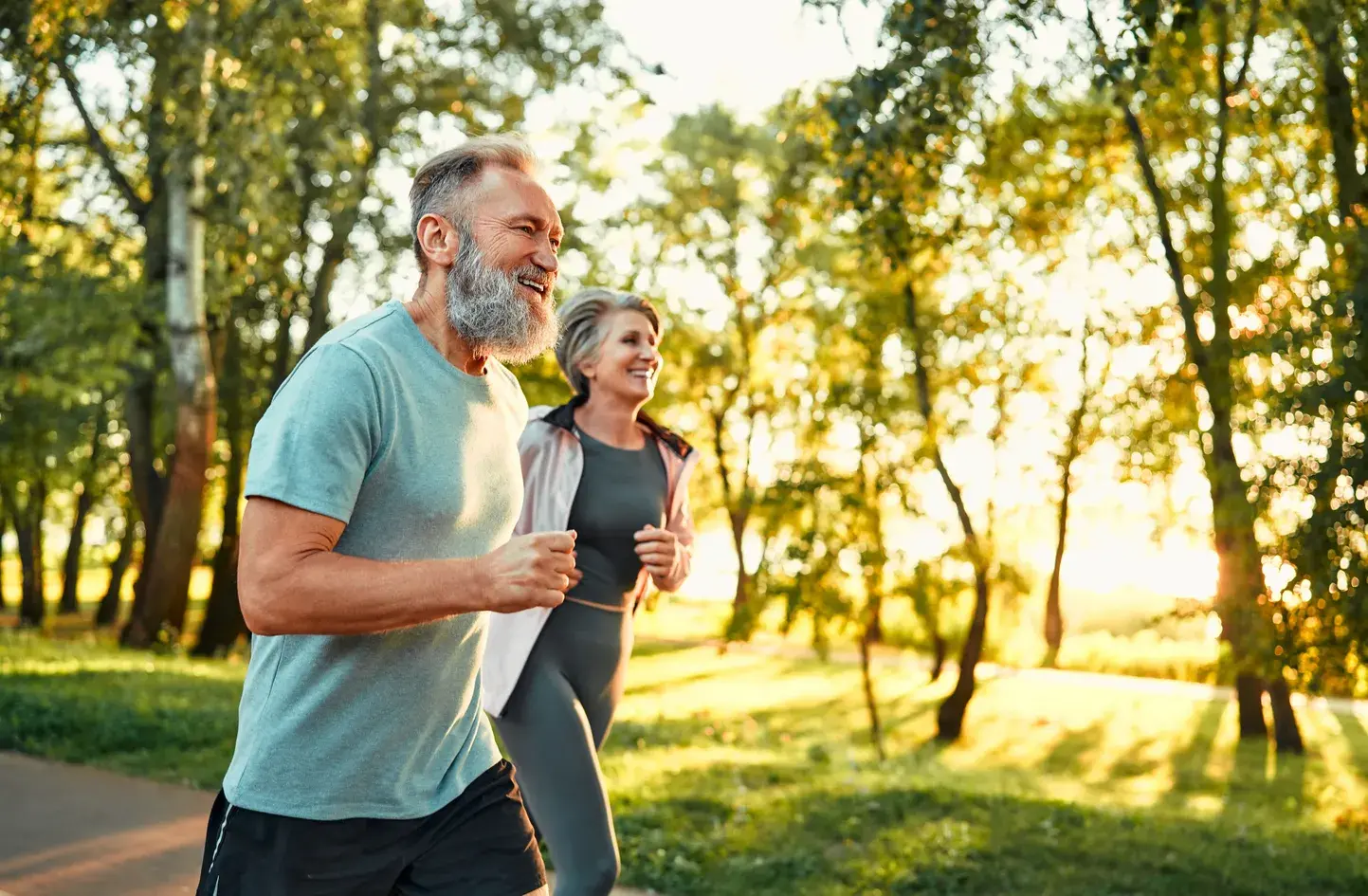Smiling elderly man and woman jogging together in a sunlit park with green trees.
