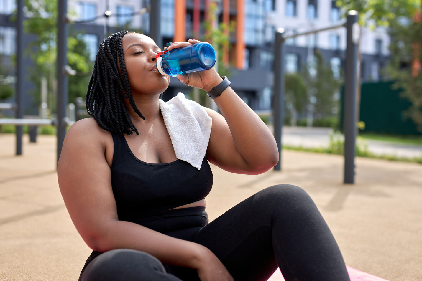 Woman in black workout clothes sitting outdoors, drinking water from a blue bottle with a white towel on her shoulder.