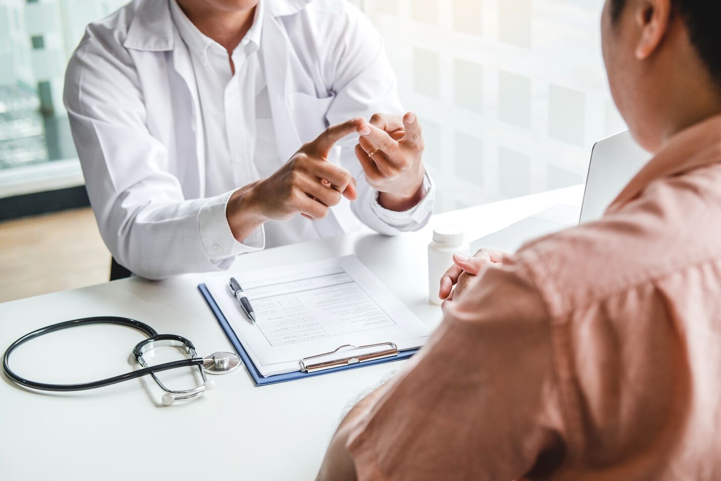 Doctor explaining a medical condition to a patient at a desk with a stethoscope, clipboard, pen, medicine bottle, and laptop.