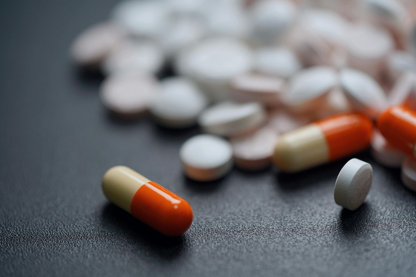 Close-up of orange and cream capsules with various white and pink round pills scattered on a dark textured surface.