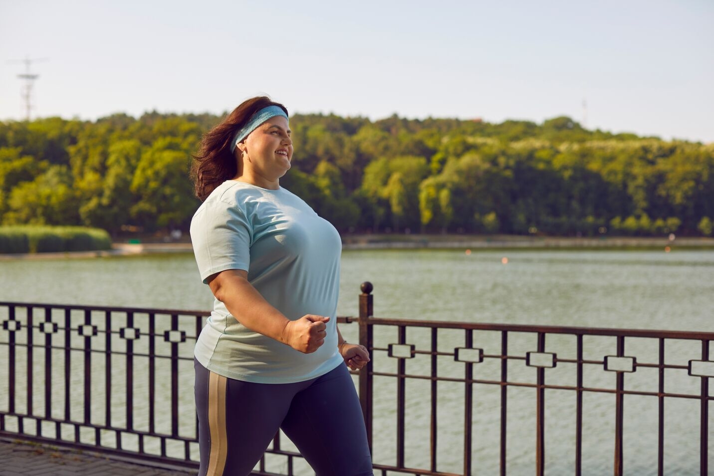 Woman walking and exercising outdoors along a railing by a river with trees in the background.