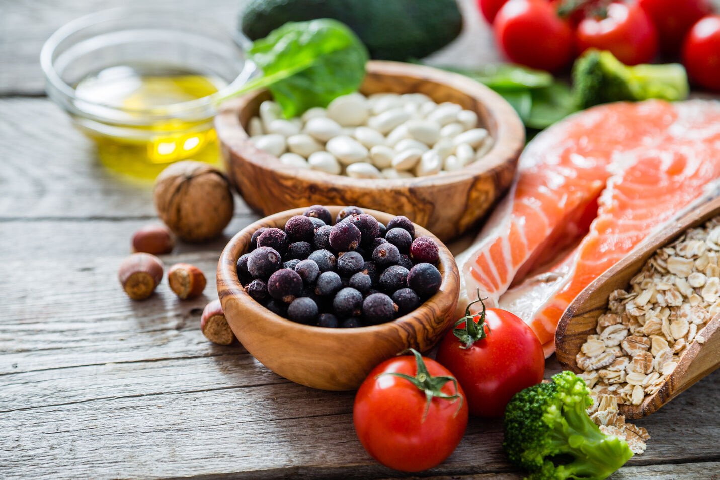 Assortment of healthy foods including blueberries, white beans, salmon, tomatoes, broccoli, oats, nuts, and olive oil on a wooden surface.