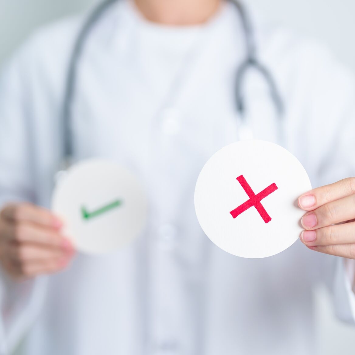 Close-up of a person in a white coat with a stethoscope holding circular cards showing a green check mark and a red X mark.