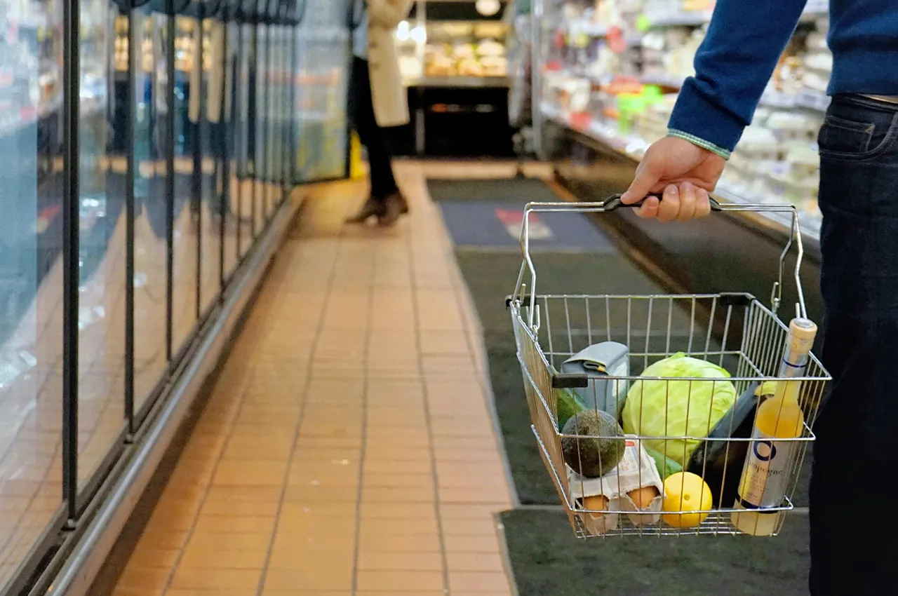 Person in blue sweater carrying a metal shopping basket with groceries in a store aisle.