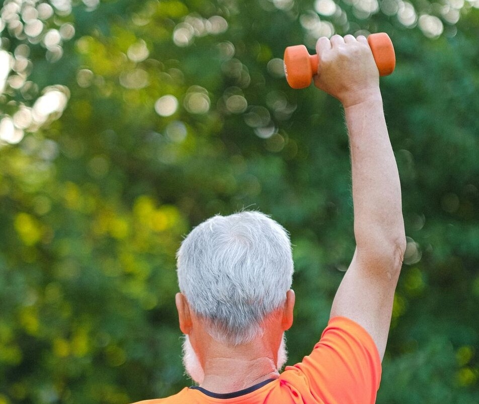 Older man with gray hair lifting an orange dumbbell overhead outdoors against a green leafy background.