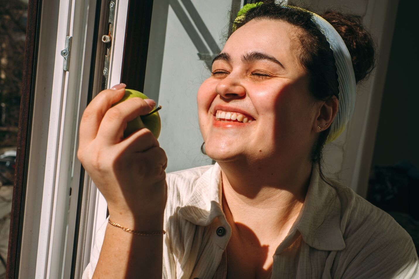 Smiling woman holding a green apple near a sunlit window.