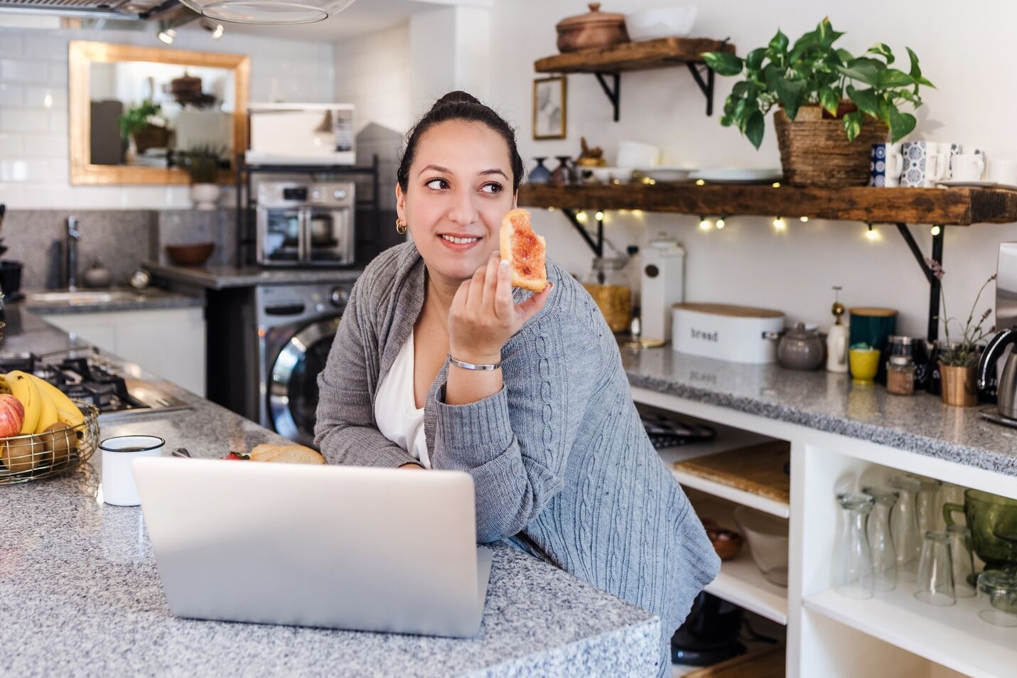 Woman in a gray cardigan eating toast with jam while sitting at a kitchen counter with a laptop.
