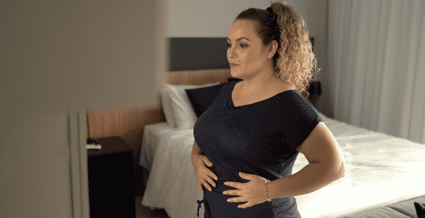 Woman with curly hair in a ponytail standing in a bedroom with her hands on her stomach.