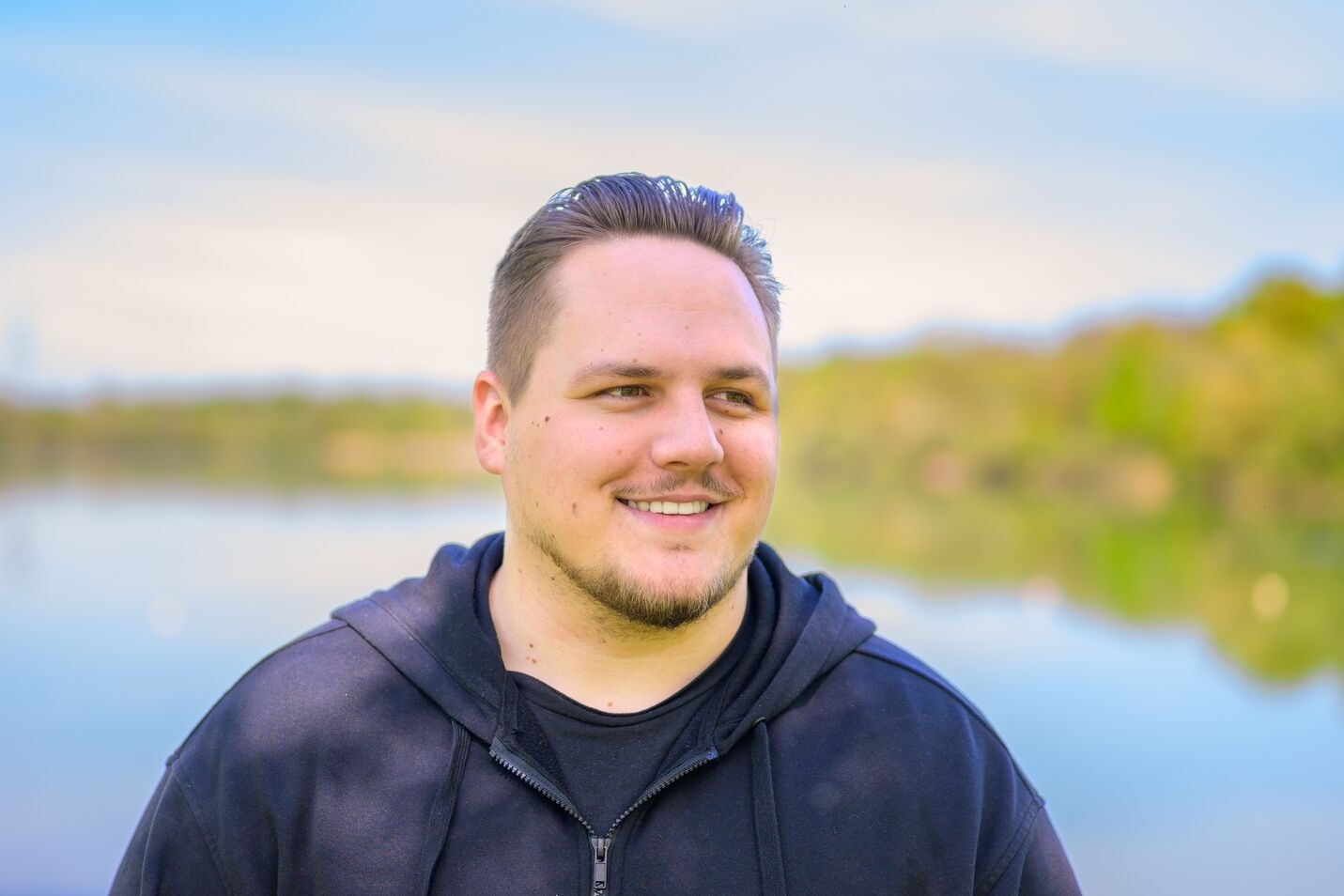 Smiling man with short dark hair and a goatee wearing a black hoodie standing outdoors near a body of water with greenery in the background.