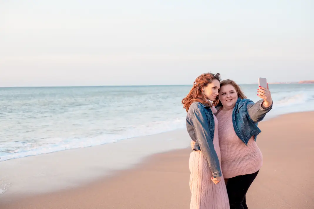 Two girls on the beach taking a photo together. 