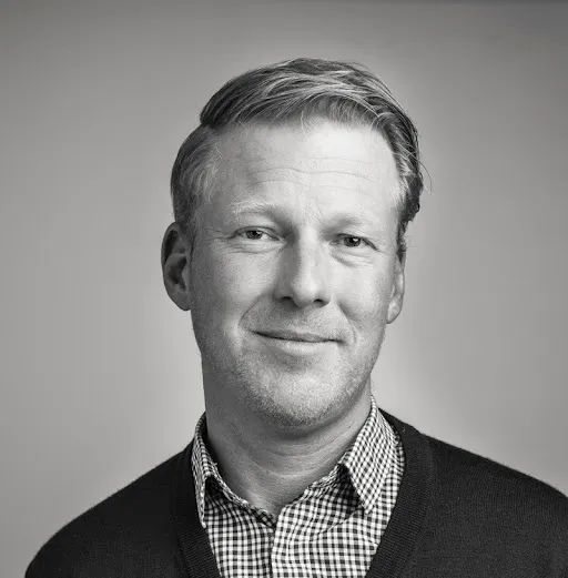 Black and white portrait of a man with short hair wearing a checkered shirt and a dark sweater, smiling slightly.