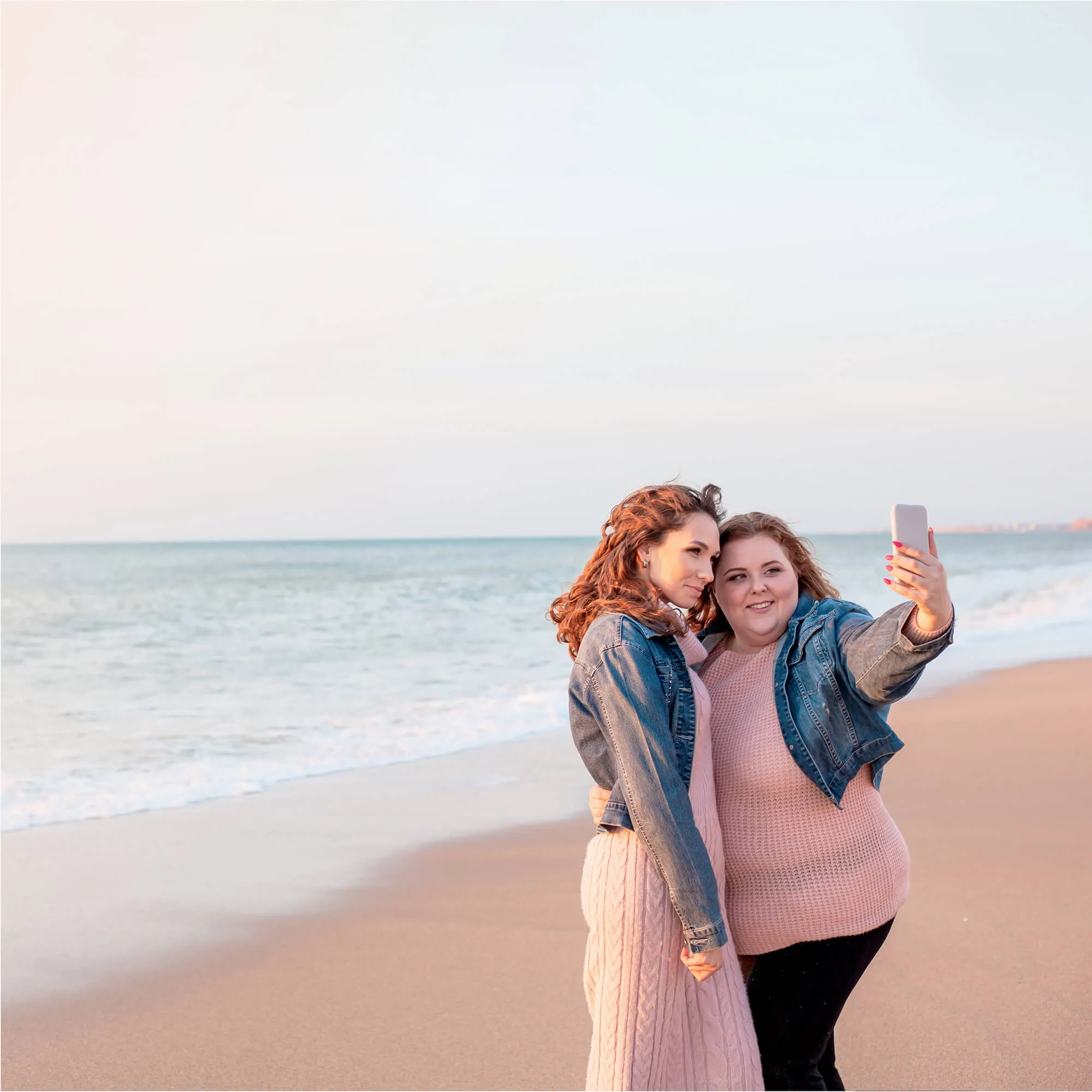 Two women in denim jackets taking a selfie together on a sandy beach near the ocean at sunset.