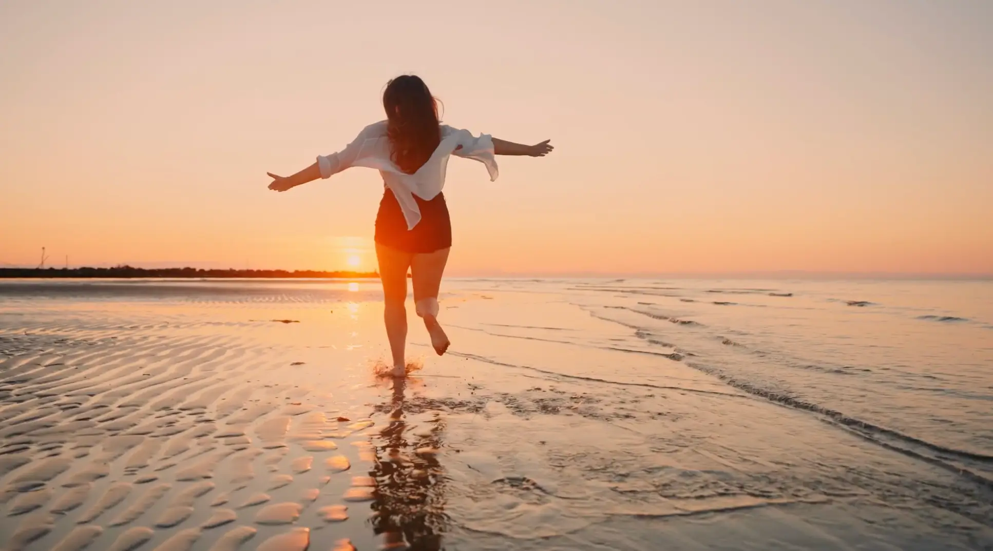 Woman running barefoot on the wet sandy beach toward the sunset with arms outstretched.