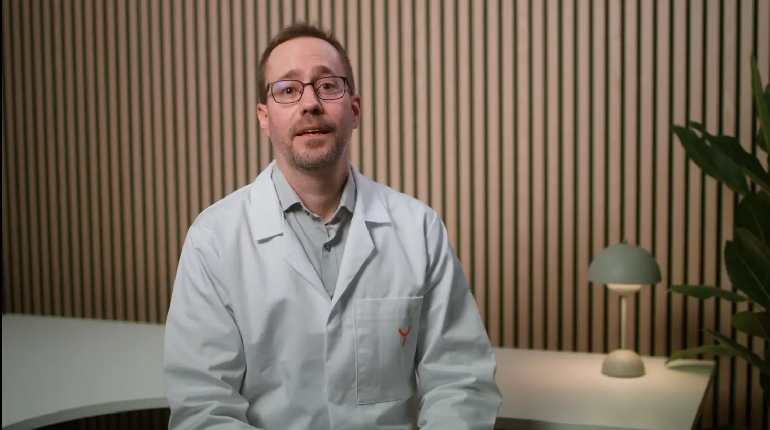 Man wearing glasses and a white lab coat sitting in front of a wooden slat wall with a modern lamp and plant in the background.
