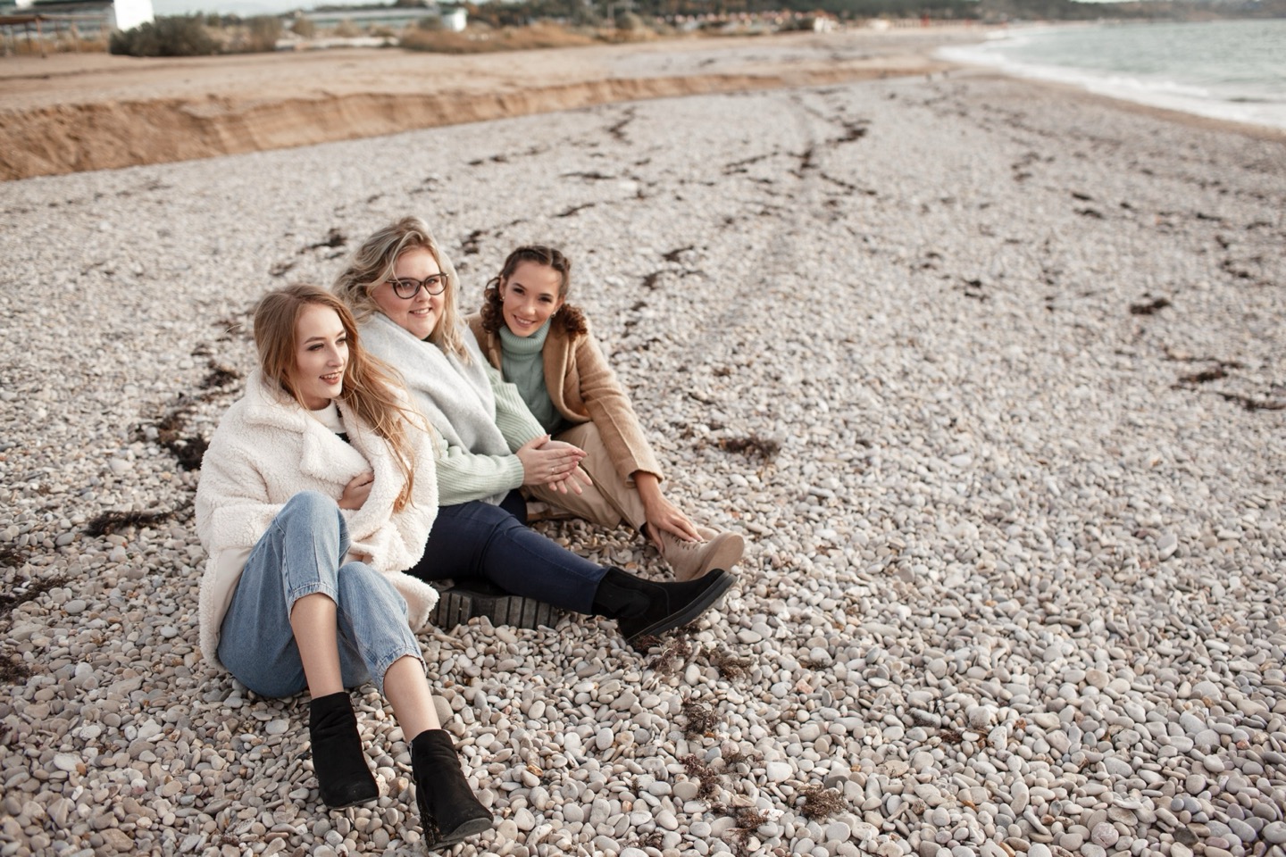 Three women sitting happily on a pebble beach near the water, dressed in warm clothing.