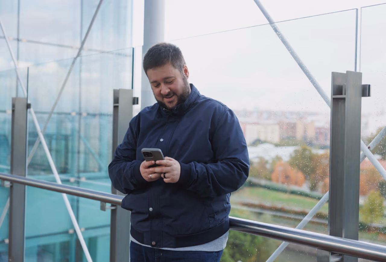 Man in navy jacket smiling while using smartphone, leaning on railing by glass wall with a city view.