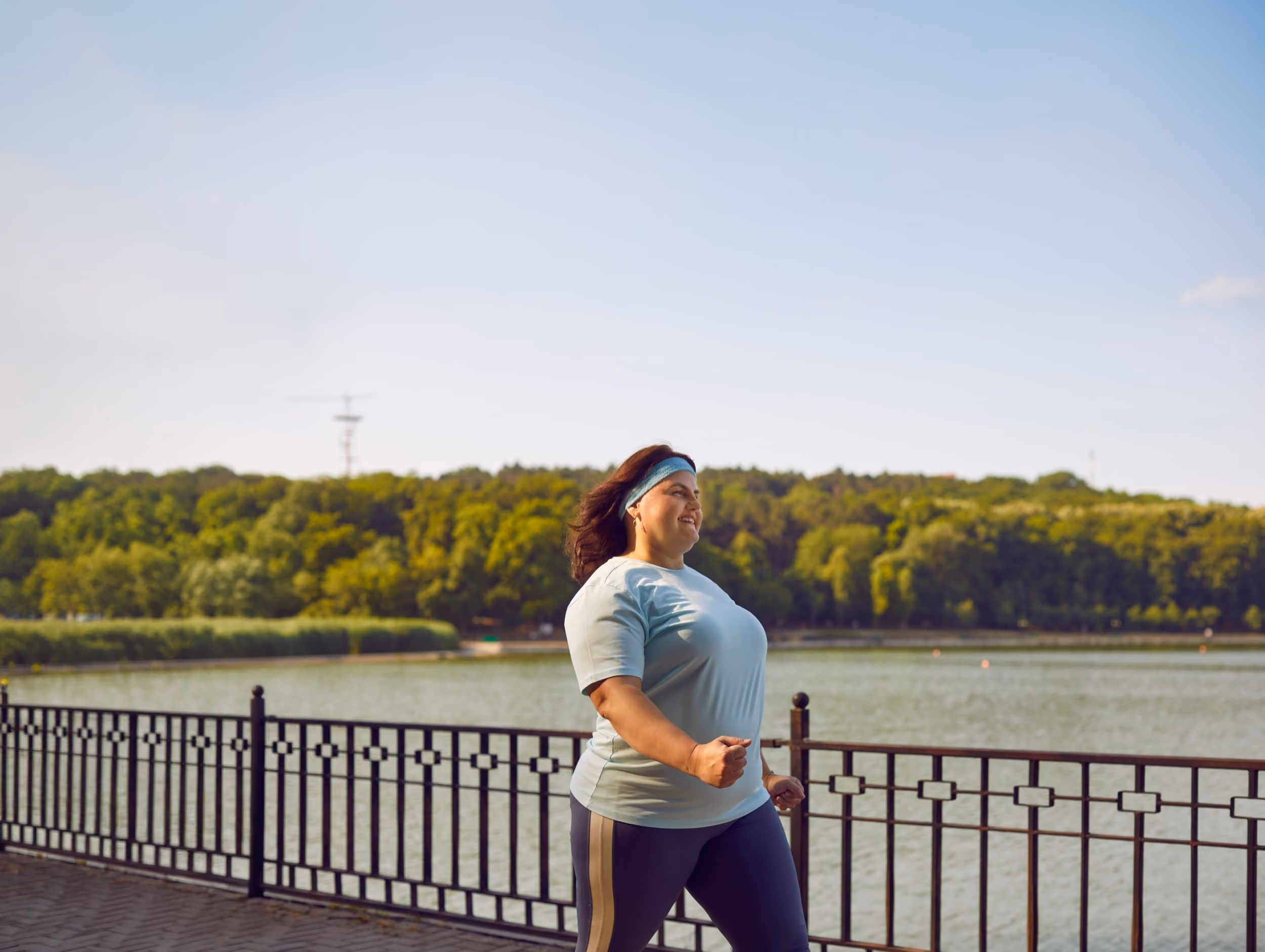 Woman jogging by a lakeside with a railing and green forest in the background on a clear day.