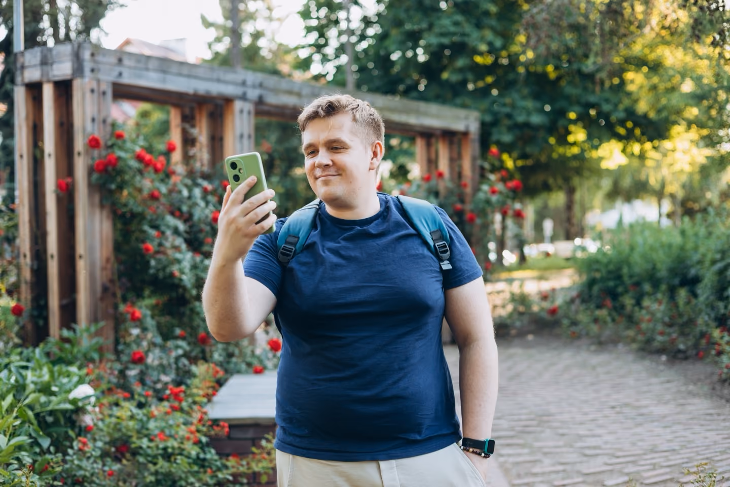 Smiling man with backpack looking at a green smartphone in a garden with red flowers.