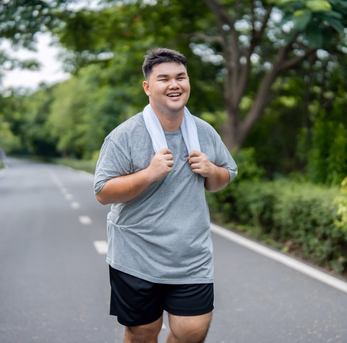 Smiling man in grey shirt with towel around neck walking on a tree-lined road.