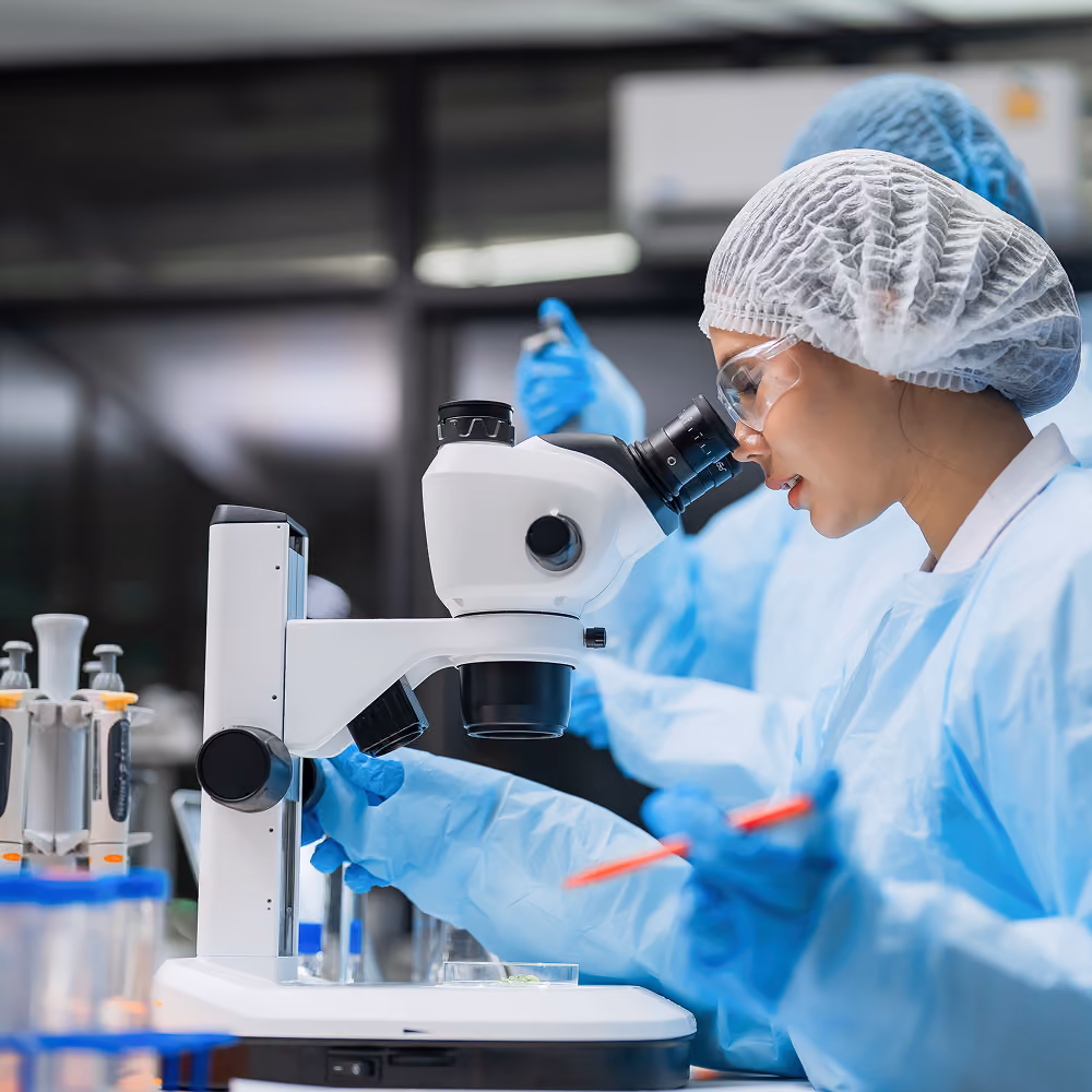 Lab technician in protective gear and gloves examining a sample under a microscope in a pharmaceutical laboratory