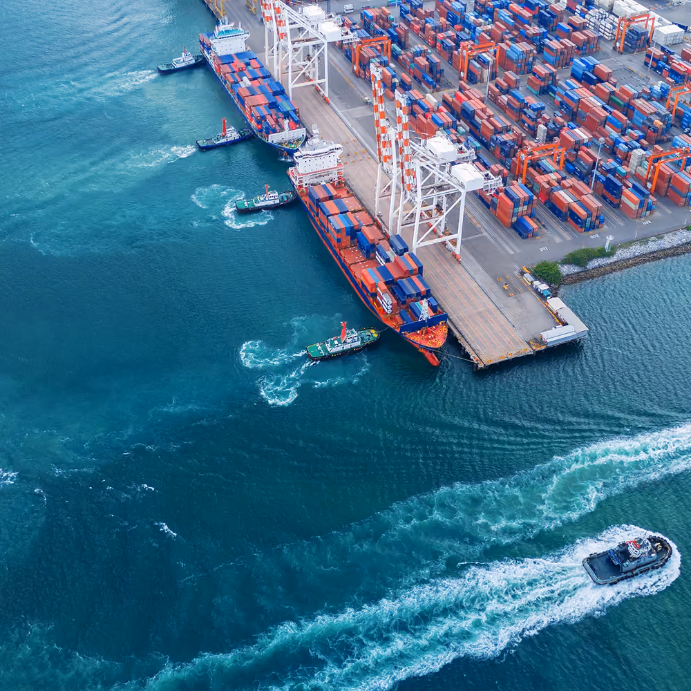 Aerial view of container ships and tugboats at a busy shipping port, with rows of cargo containers along the dock