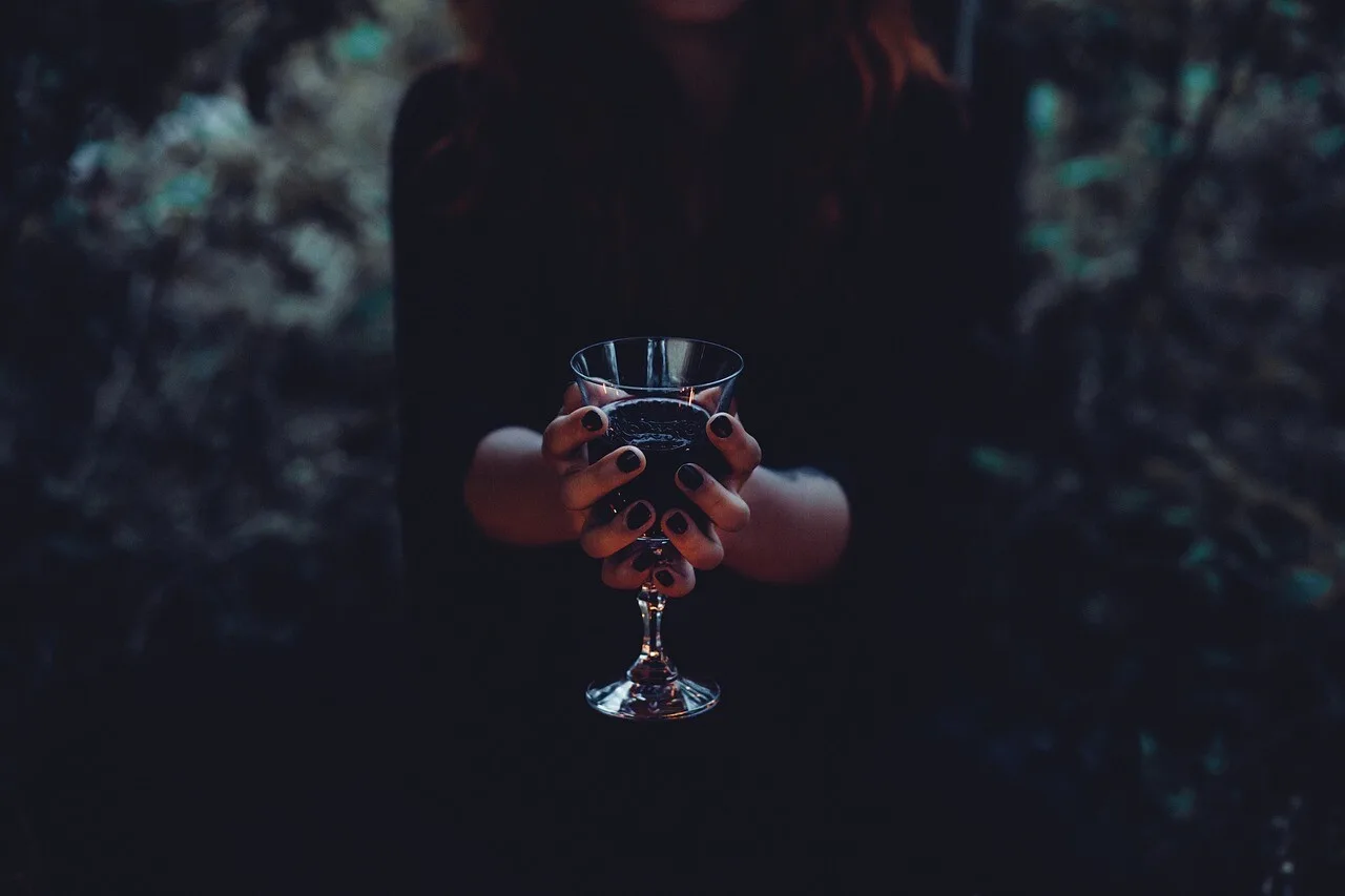 Hands holding up a coupe glass of wine in dim light among dark foliage