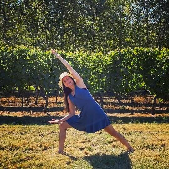 A woman in a sun hat holding a yoga warrior pose in front of the vineyard rows at 14 Acres