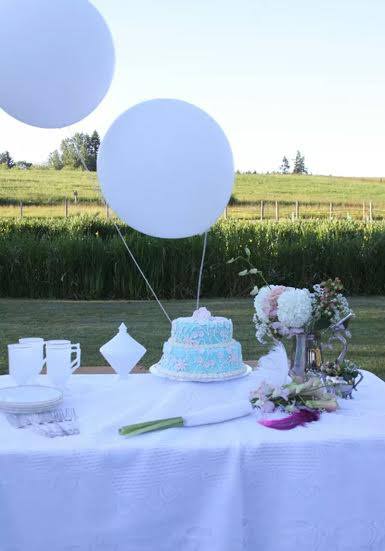 A birthday cake table with white balloons and flowers set up on the lawn beside the vineyard