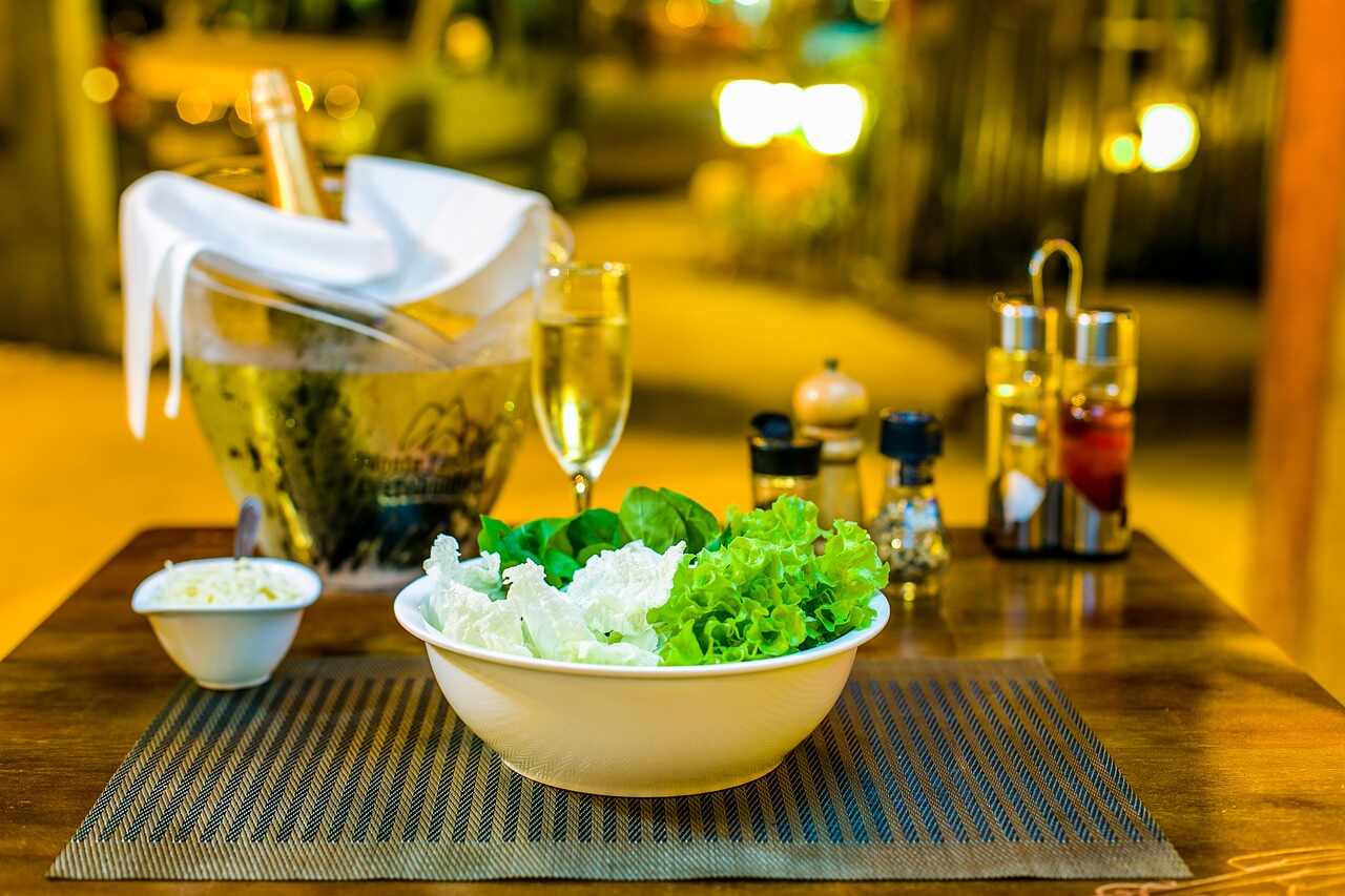 A bottle of sparkling wine in an ice bucket beside a glass and a bowl of green salad