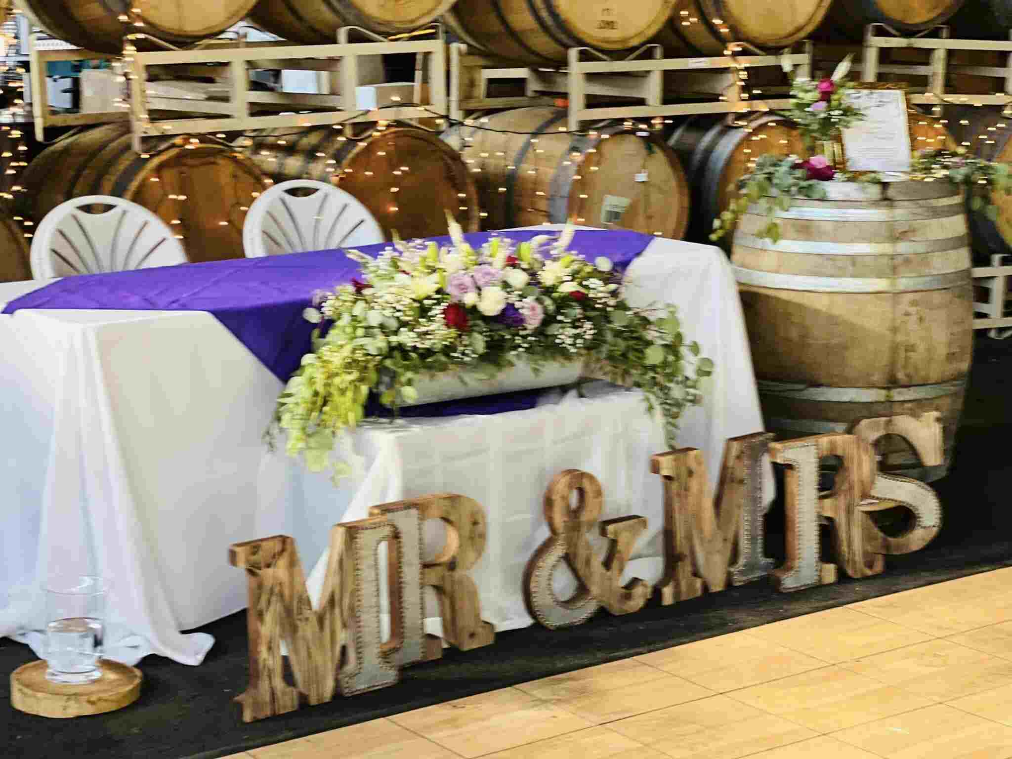 A sweetheart table with white linen, a purple runner, floral garland and wooden MR & MRS letters in the barrel room