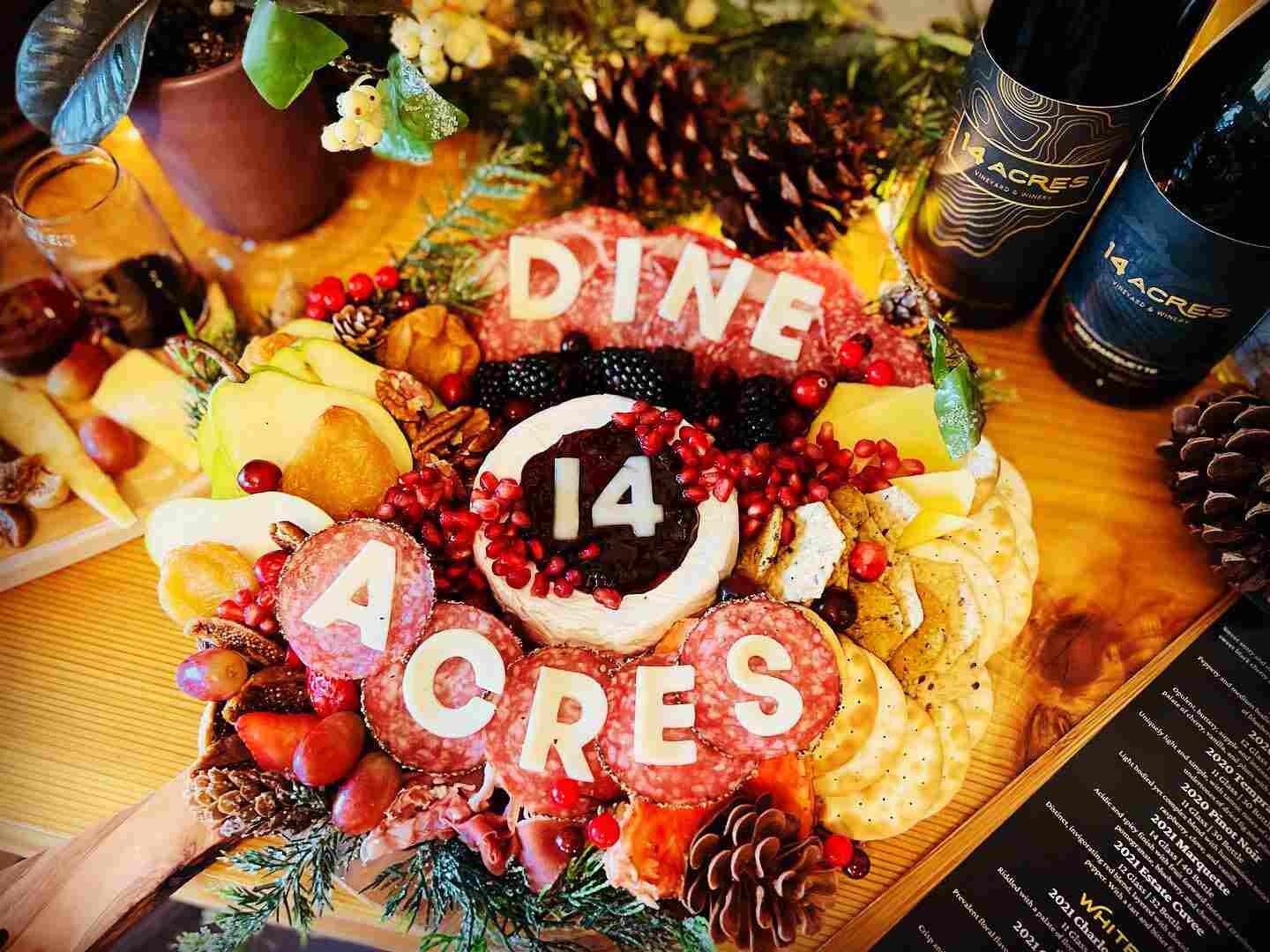 A holiday charcuterie board spelling out DINE 14 ACRES in cheese and fruit, with two bottles of 14 Acres wine
