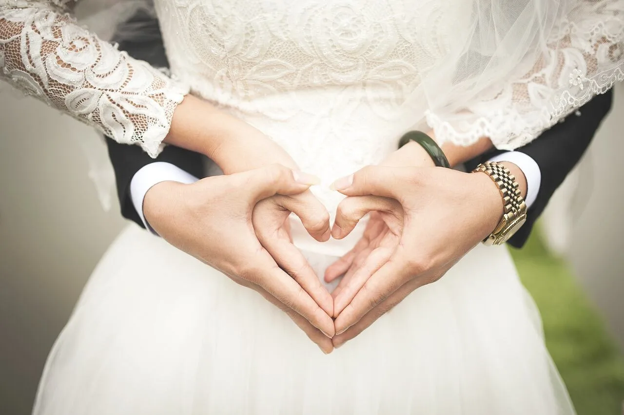 A bride and groom's hands forming a heart in front of her white lace wedding dress