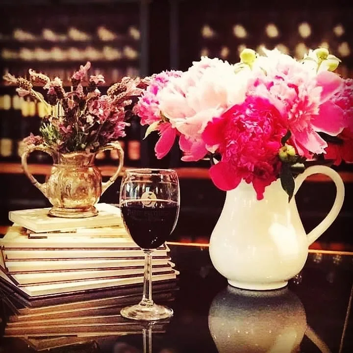 A vase of pink peonies and a glass of red wine on a table in front of the wine racks