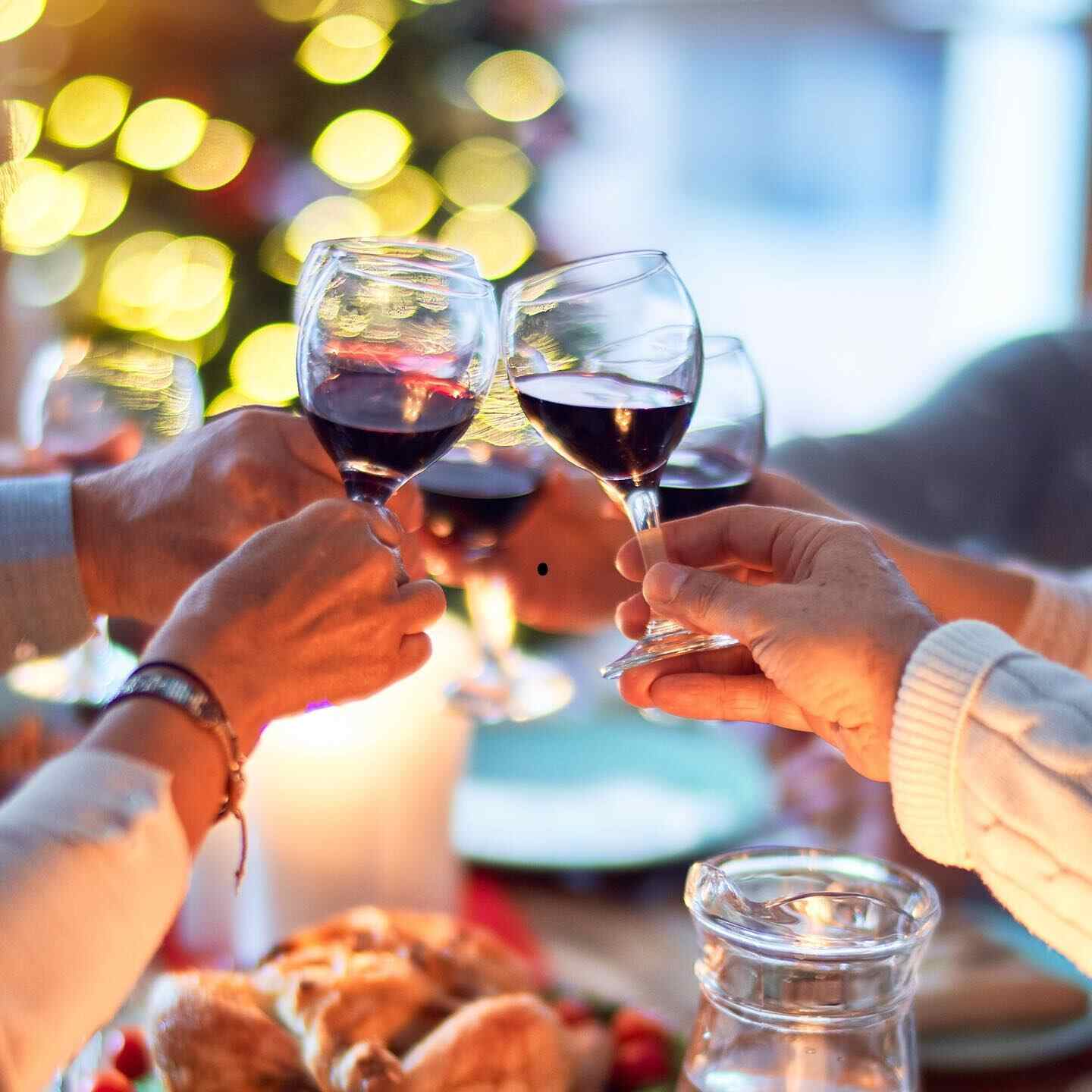 Hands clinking glasses of red wine over a table of food, warm string lights behind