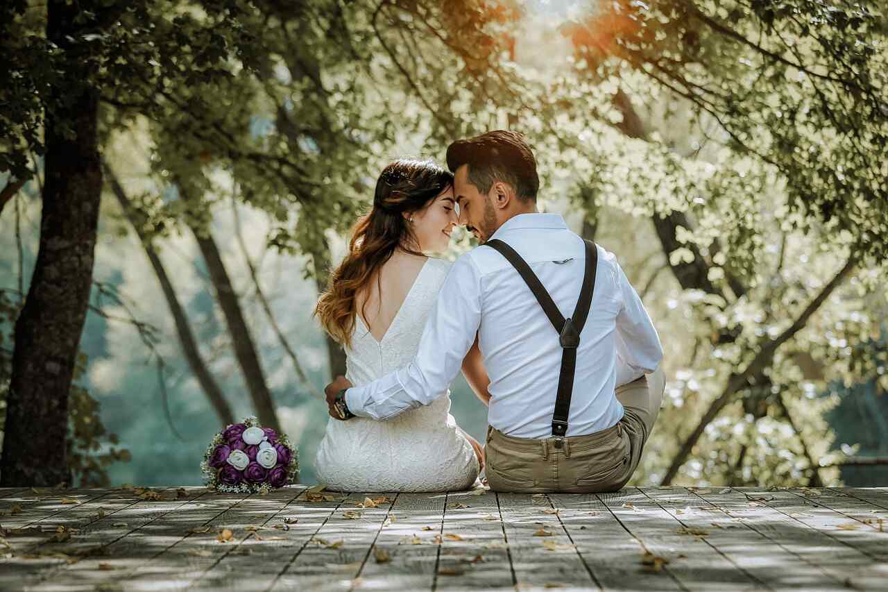A bride and groom sitting close together on a wooden deck under blossoming trees, her bouquet beside them