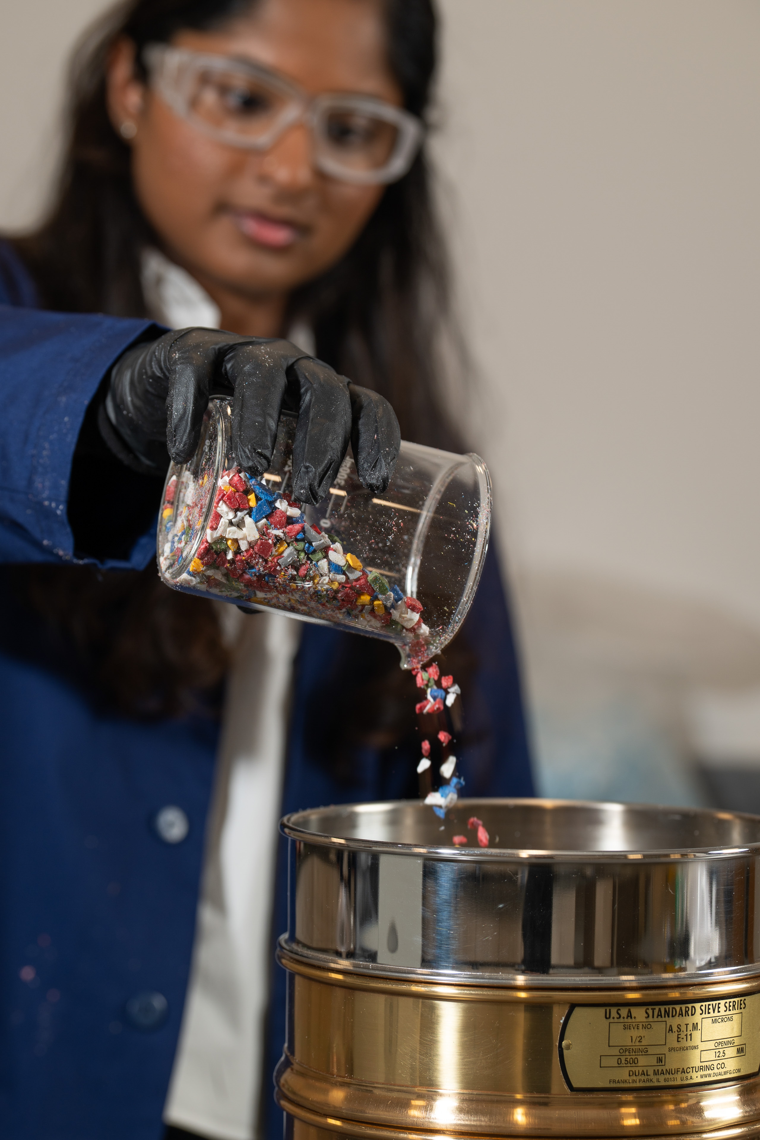 Lab technician in safety glasses and black gloves pouring color-coded plastic flakes into a testing sieve