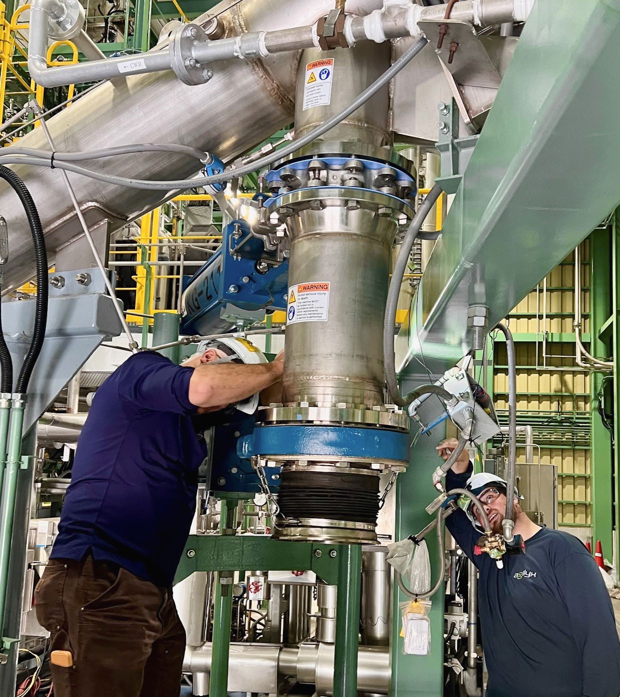 Two Agilyx technicians in hard hats servicing a large steel reactor vessel with blue flange fittings at the recycling plant