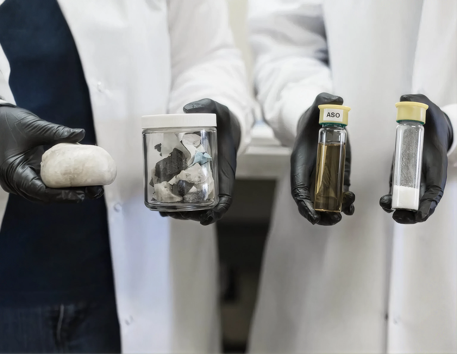 Lab researchers in white coats and black gloves holding samples of recycled plastic feedstock and processed liquid outputs