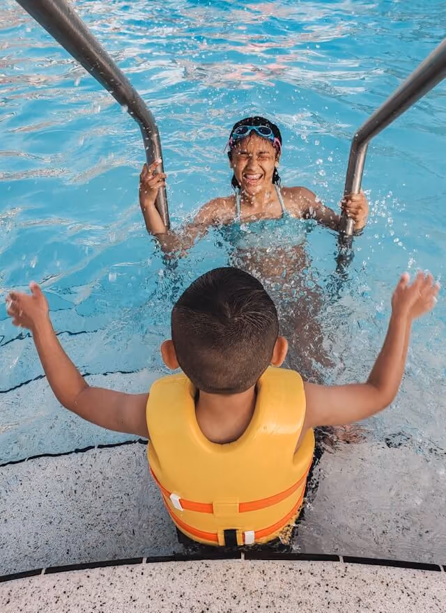 Two kids playing in the shallow end, one looking up smiling at the camera.