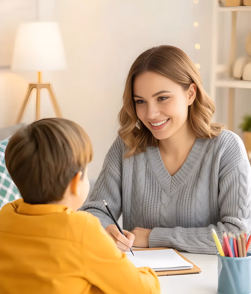 Woman smiling and talking to a child while holding a pencil over a notebook in a cozy, well-lit room.