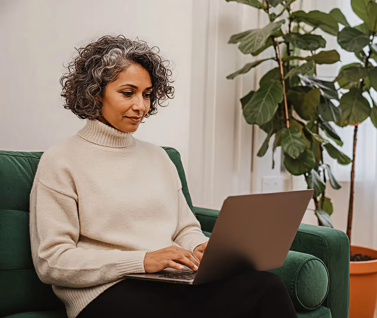 Middle-aged woman with curly salt-and-pepper hair wearing a beige turtleneck sweater, sitting on a green couch and working on a laptop.
