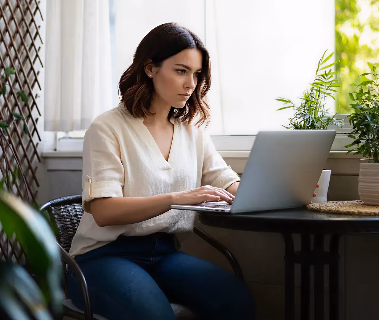 Woman with short dark hair wearing a white blouse working on a laptop at a table near a window with plants.