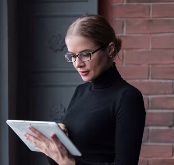 Woman with glasses and a black turtleneck using a white tablet device against a brick wall background.