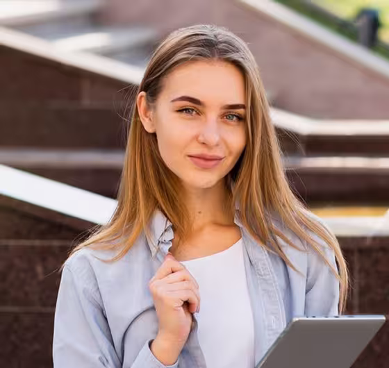 Young woman with long blonde hair holding a tablet outdoors, smiling with stairs in the background.