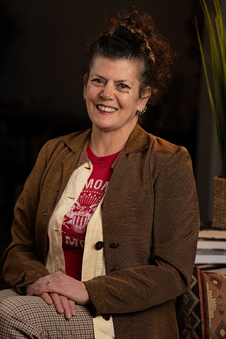 Ruby Dee Philippa. A woman smiling at the camera with curly brown hair wearing a brown jacket, red Ramones T-shirt, and gray pants.